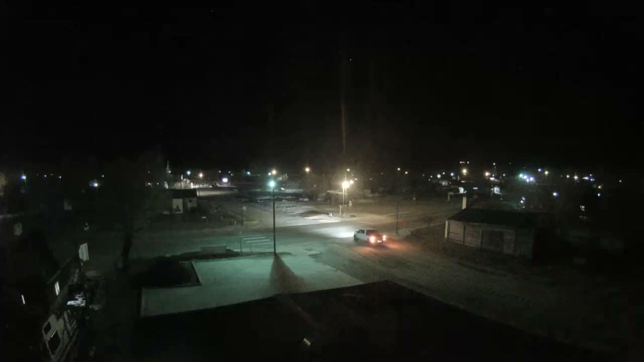 A white pickup truck with its headlights and taillights on drives through a small town at night, where streetlights illuminate roads and scattered buildings under clear, cold conditions with patches of snow or ice visible on the ground.