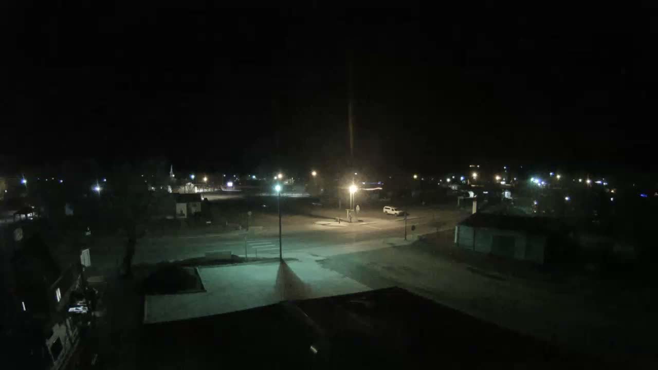 An elevated view of a small town at night shows clear weather, with streetlights illuminating an intersection and surrounding buildings, and a white vehicle parked near the road.