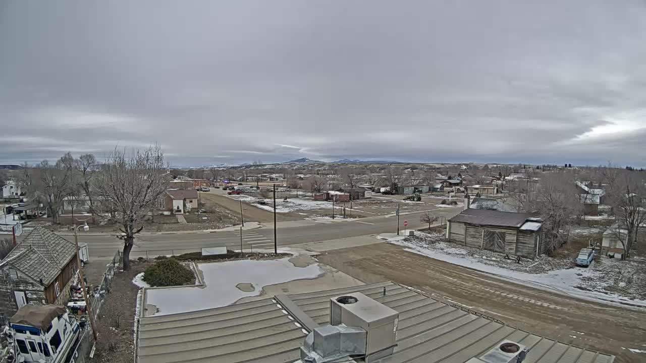 The image depicts an overcast winter day in a small town, featuring bare trees, patches of snow on the ground, and distant hills under a gray sky, as seen from a rooftop.
