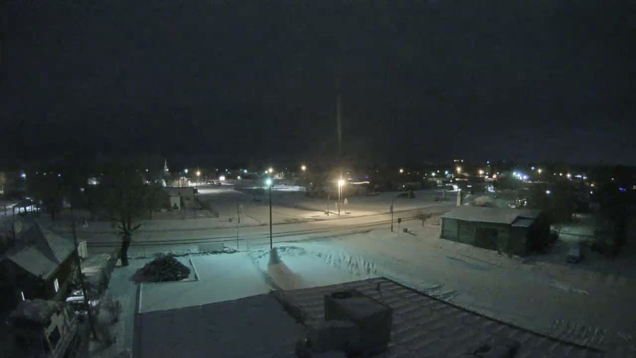 An elevated view captures a snow-covered town at night, with streetlights illuminating the freshly fallen snow on roads and rooftops under cold, clear skies.
