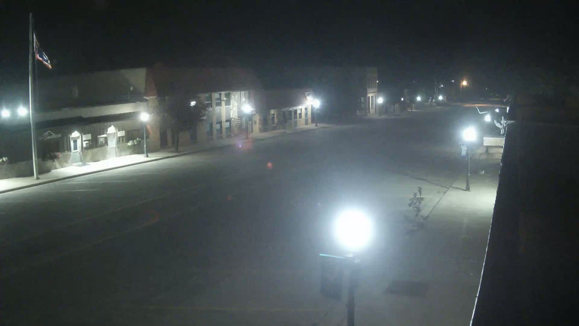 A mostly empty street at night, lined with buildings, is illuminated by streetlights.