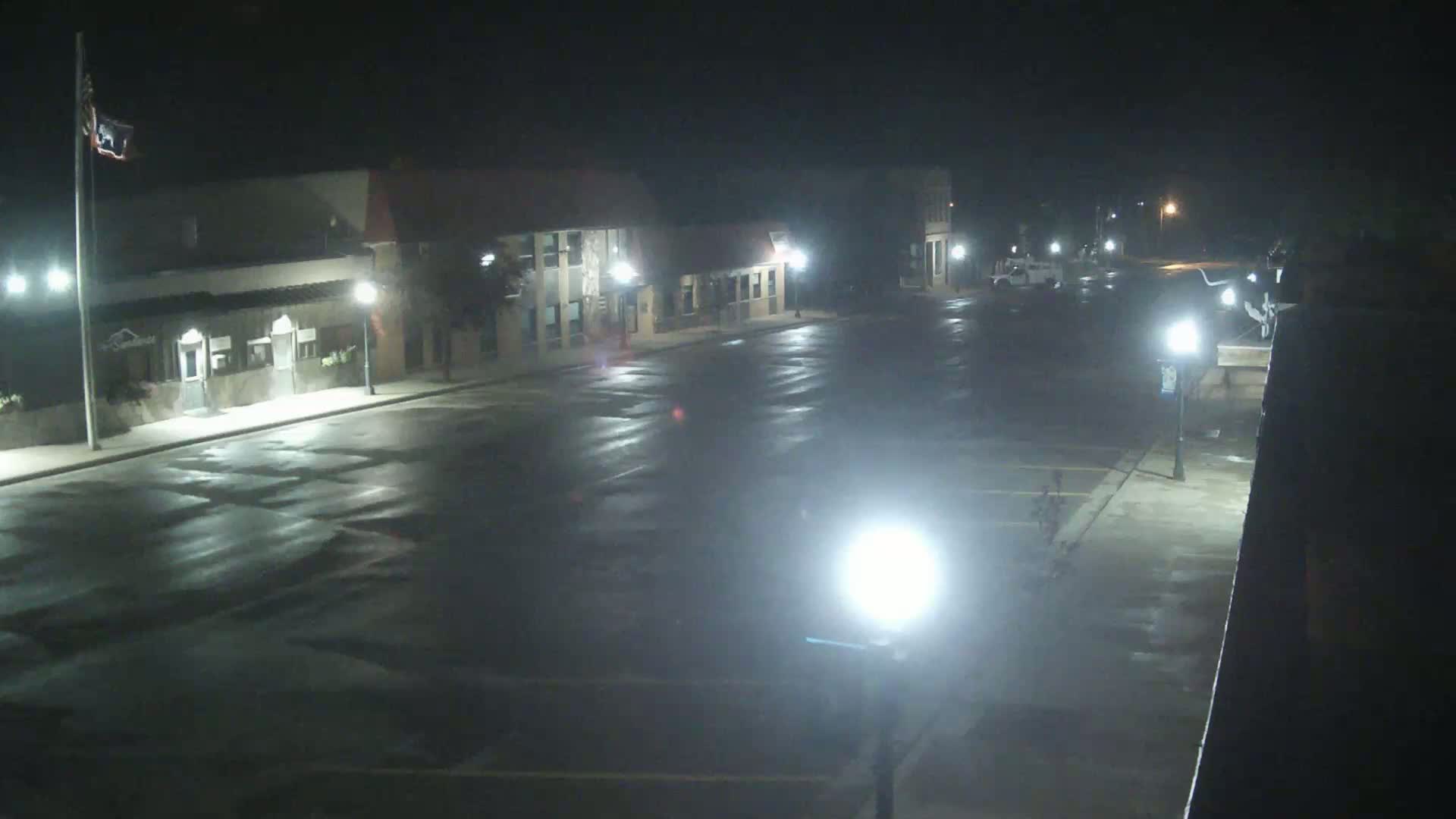 A wet, empty street at night is lit by several streetlights, with buildings lining either side.