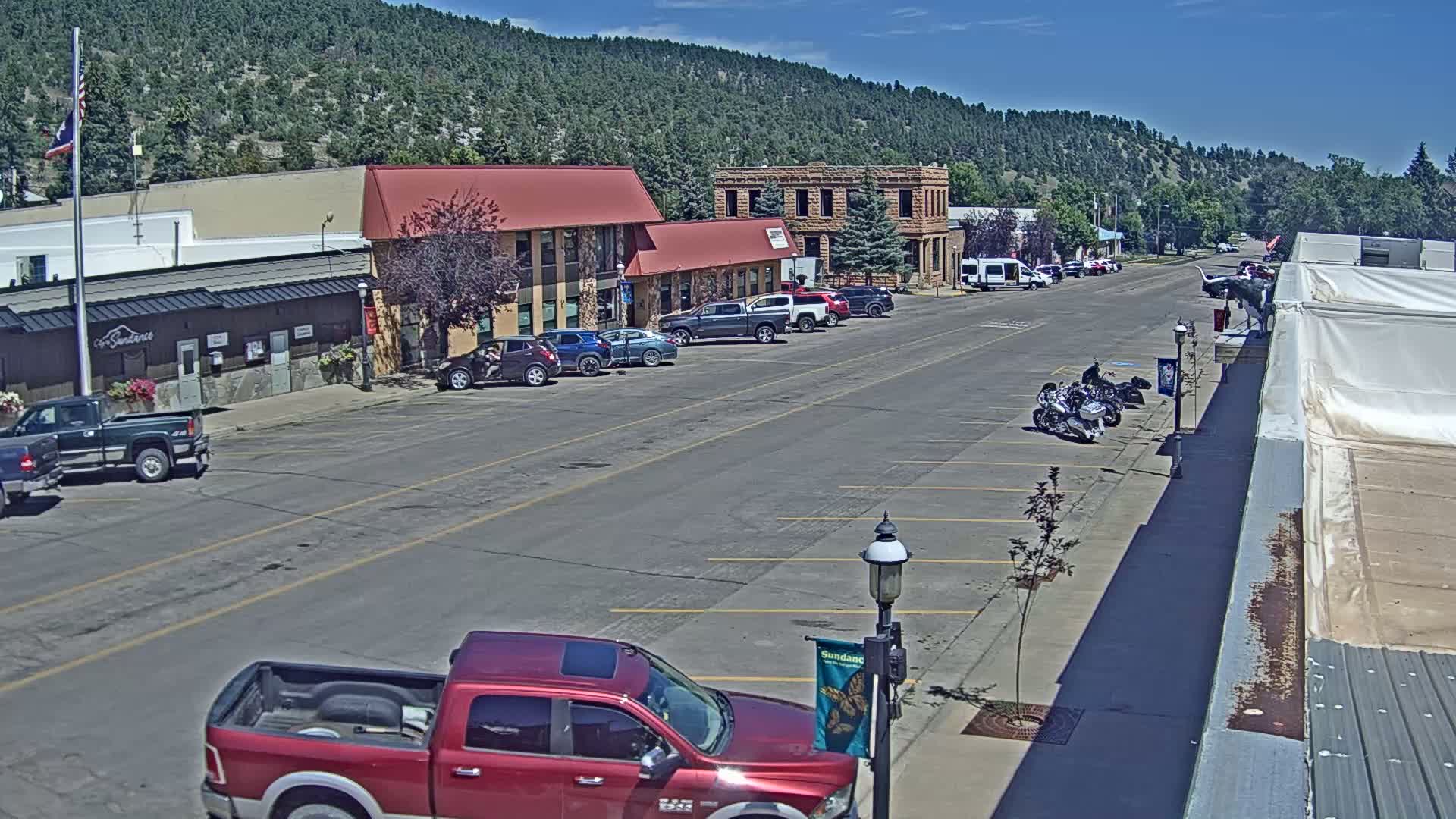 A mostly empty street in a small town, lined with parked cars and a few motorcycles, is nestled against a hillside covered in dark green trees under a clear, sunny sky.