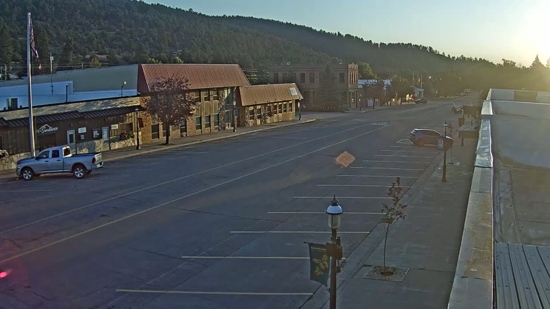 A mostly empty street in a small town, lined with parked cars and a few motorcycles, is nestled against a hillside covered in dark green trees under a clear, sunny sky.