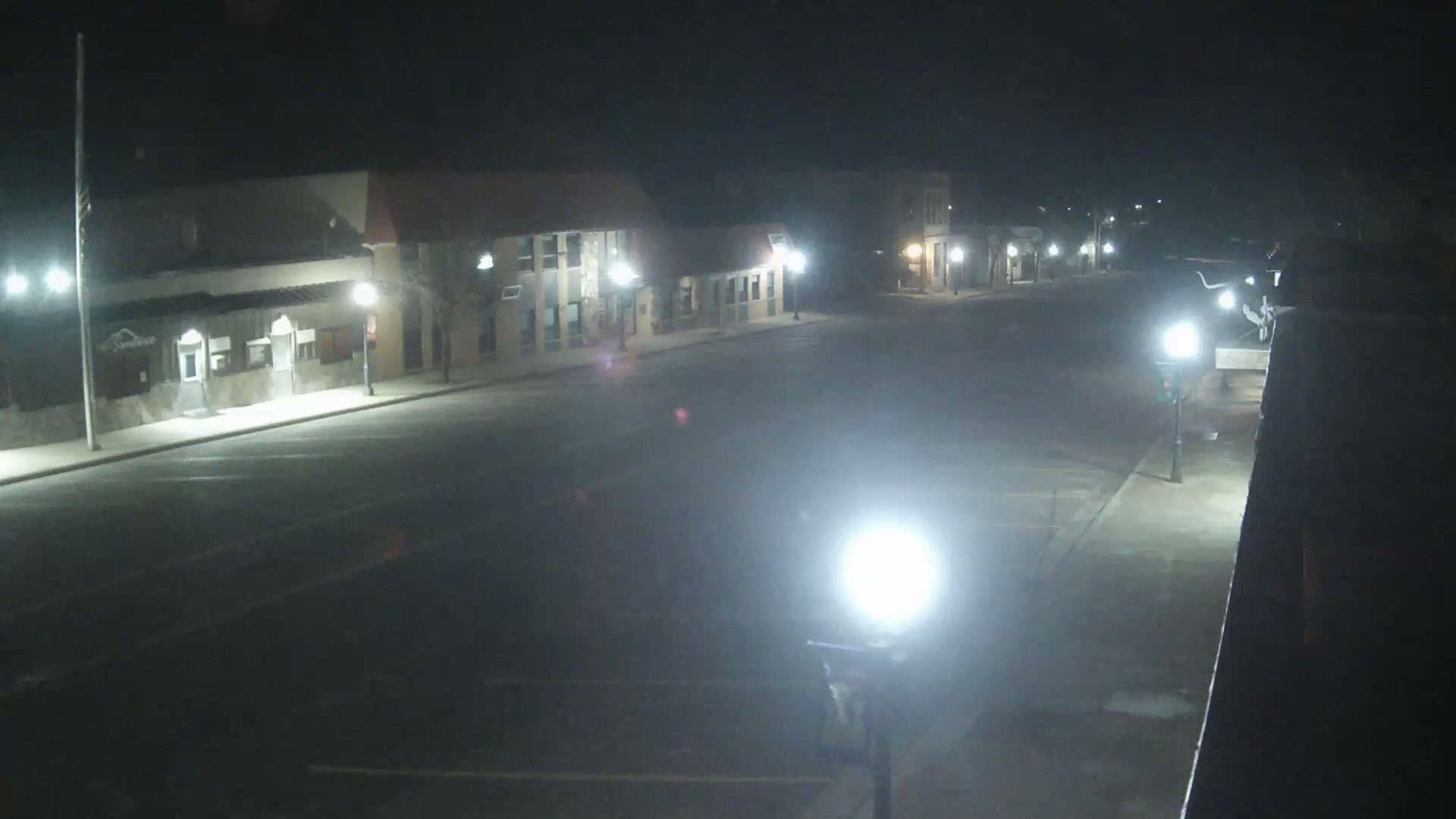 An empty town street with modest buildings is brightly lit by streetlights and distant vehicle headlights on a clear, damp night.