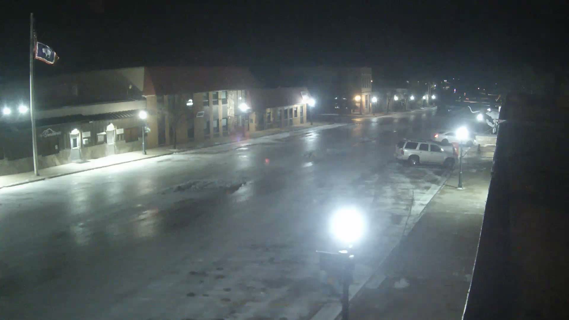A deserted street is illuminated by bright streetlights at night, with buildings lining both sides of the road under a clear sky, and a flag visible on a flagpole to the left.