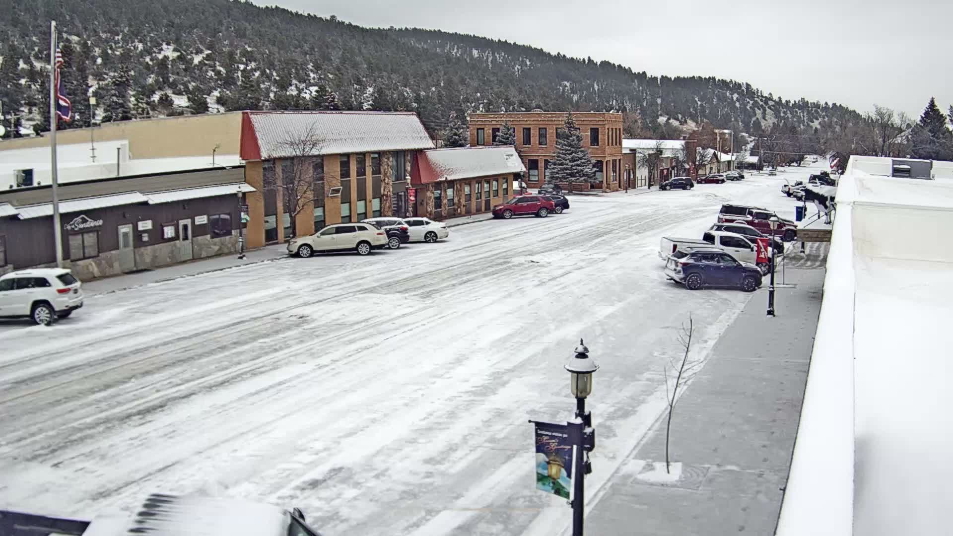 A snow-covered town street is lined with buildings and parked cars under an overcast sky, with snow-dusted hills in the background.