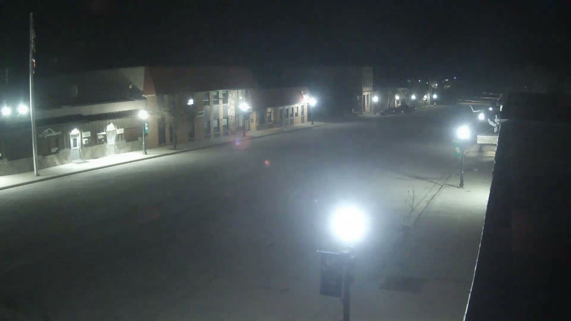 An empty street at night is illuminated by bright streetlights and the glowing windows of buildings lining both sides, with patches of ice and melted snow visible on the ground.