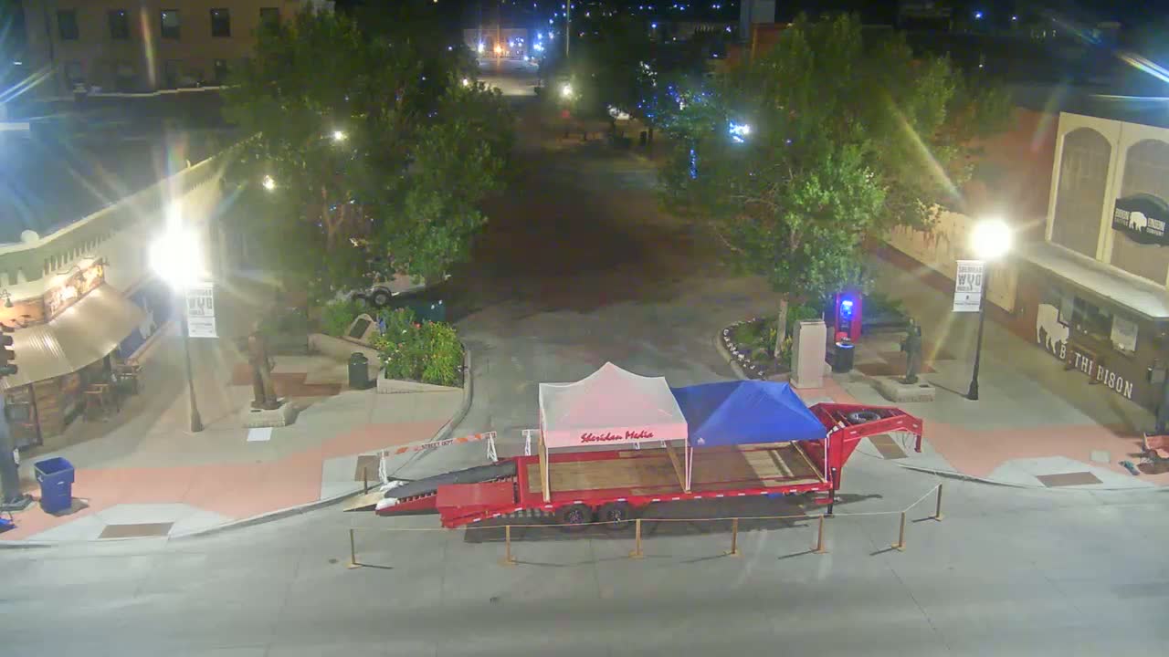 A red trailer carrying two tents sits in a mostly empty town square at night, under partly cloudy skies.