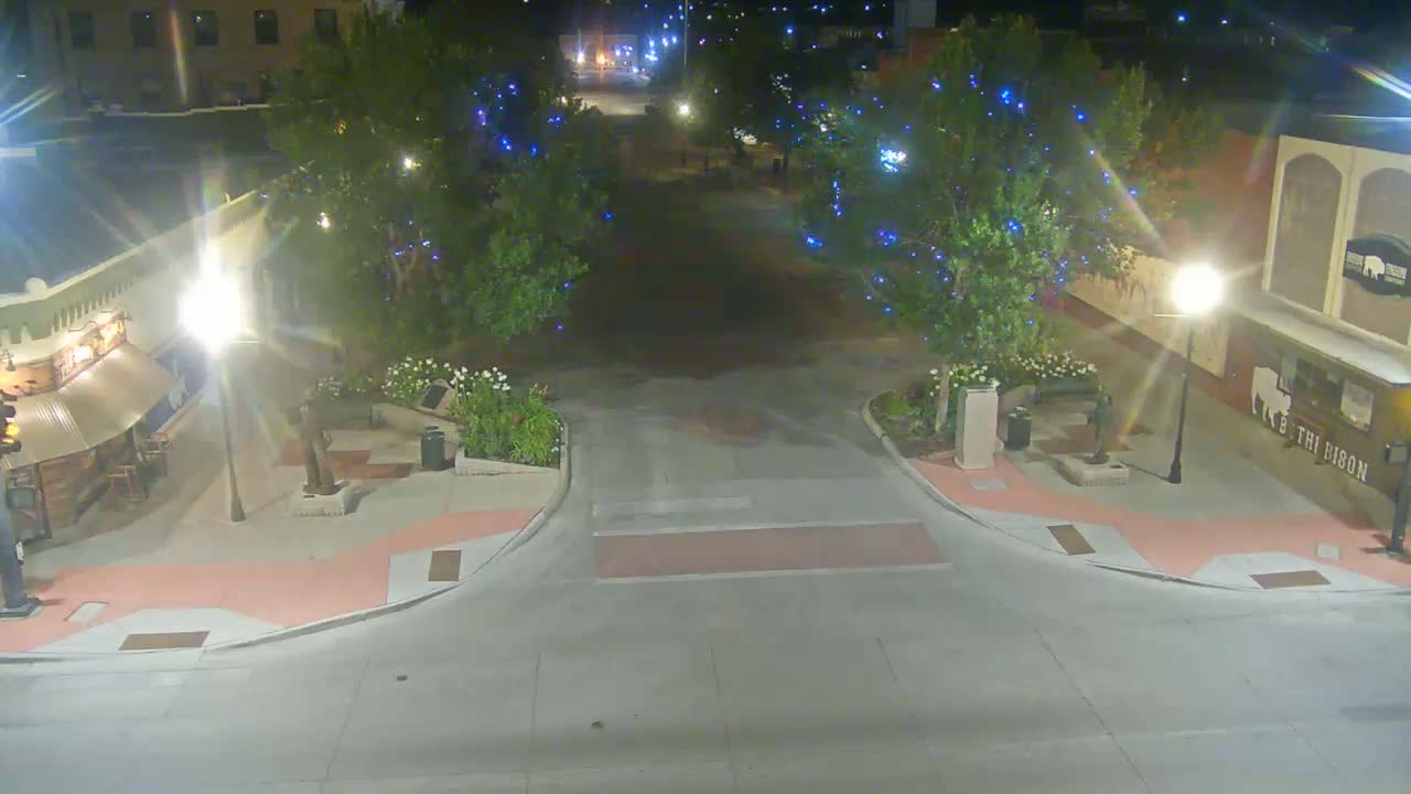 A nighttime, street-level view of a small town square with trees decorated with blue lights, streetlights, and landscaping.