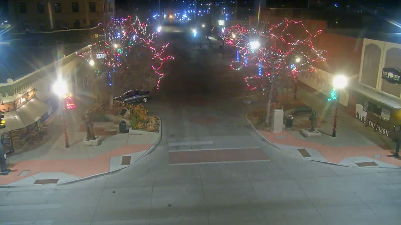 A clear night reveals a festive town square or street brightly illuminated by colorful holiday lights on trees and prominent streetlights, with surrounding buildings visible.