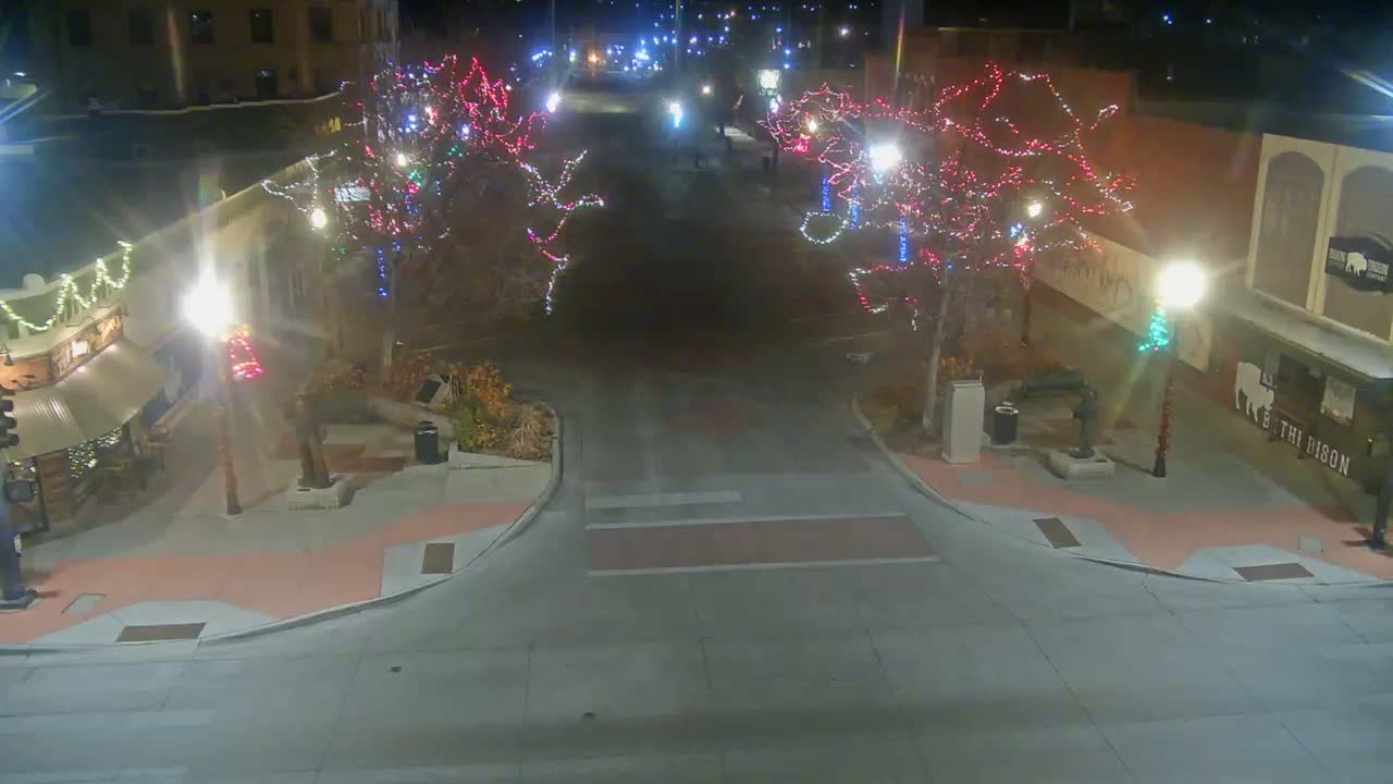 A clear night illuminates an outdoor street or plaza adorned with trees wrapped in colorful string lights, bordered by buildings with glowing windows and prominent street lamps.