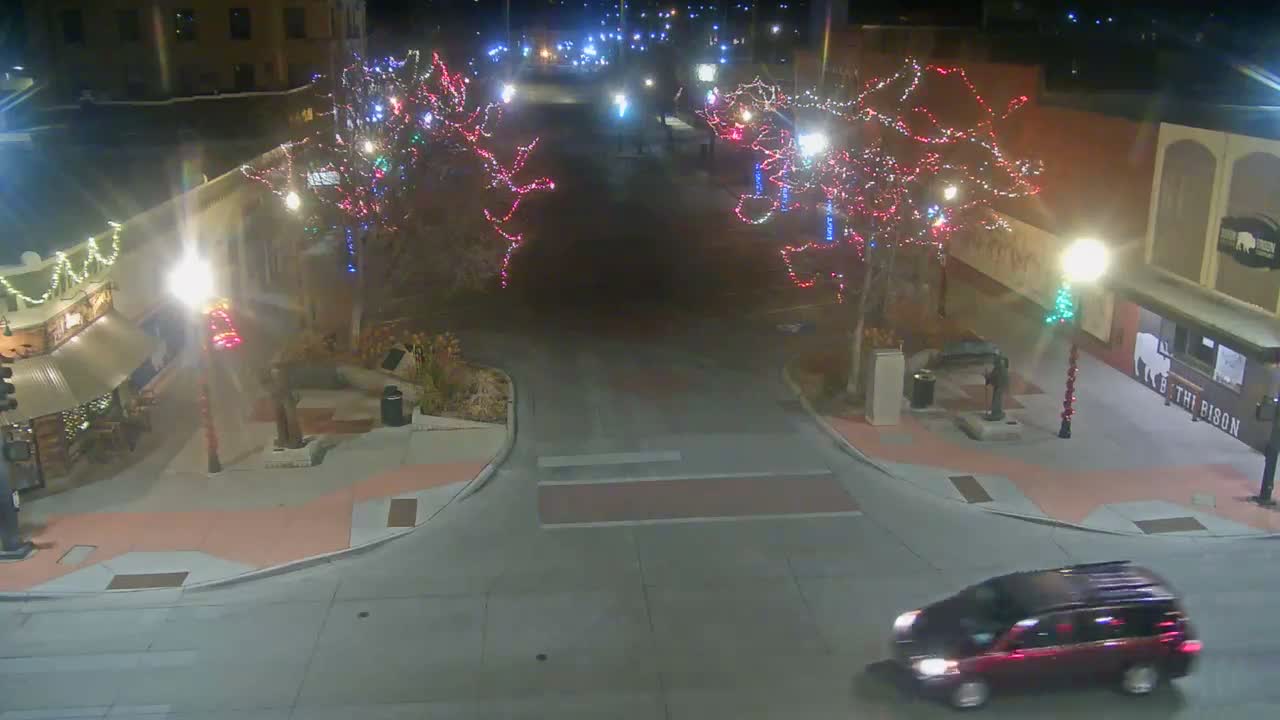 A street at night is illuminated by numerous colorful holiday lights on trees and buildings, with a red car driving in the foreground on the dry pavement under clear conditions.