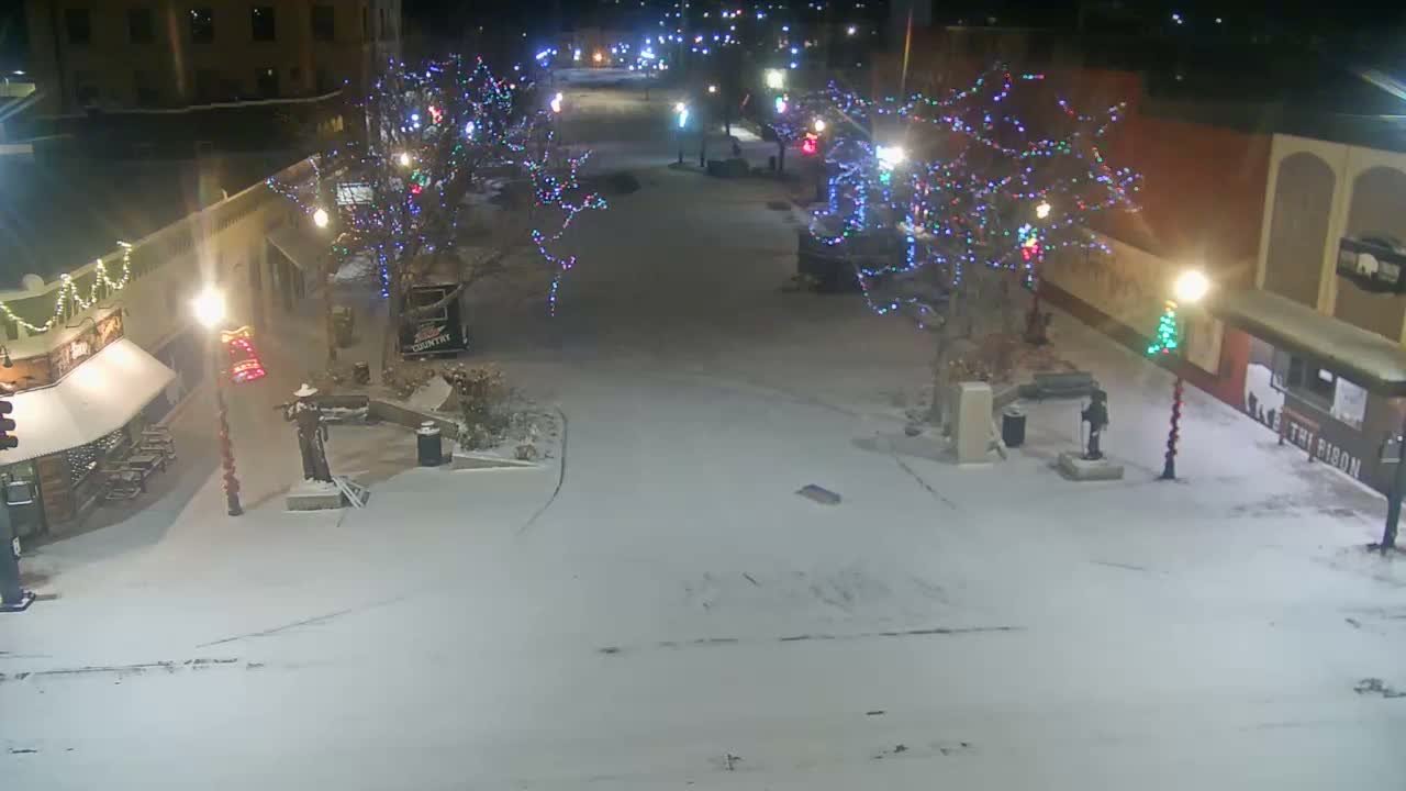 A quiet, snow-covered town street is seen at night, illuminated by numerous streetlights and festive multi-colored lights wrapped around bare trees, under cold and snowy weather.