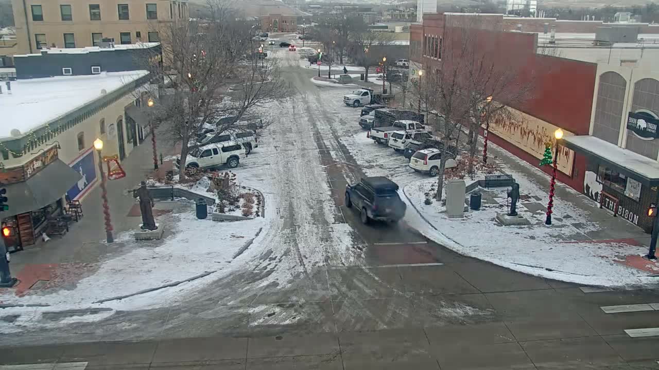 An elevated perspective captures a snowy and slushy town street lined with buildings and bare trees with holiday decorations, featuring parked cars and one moving vehicle, on an overcast winter day.
