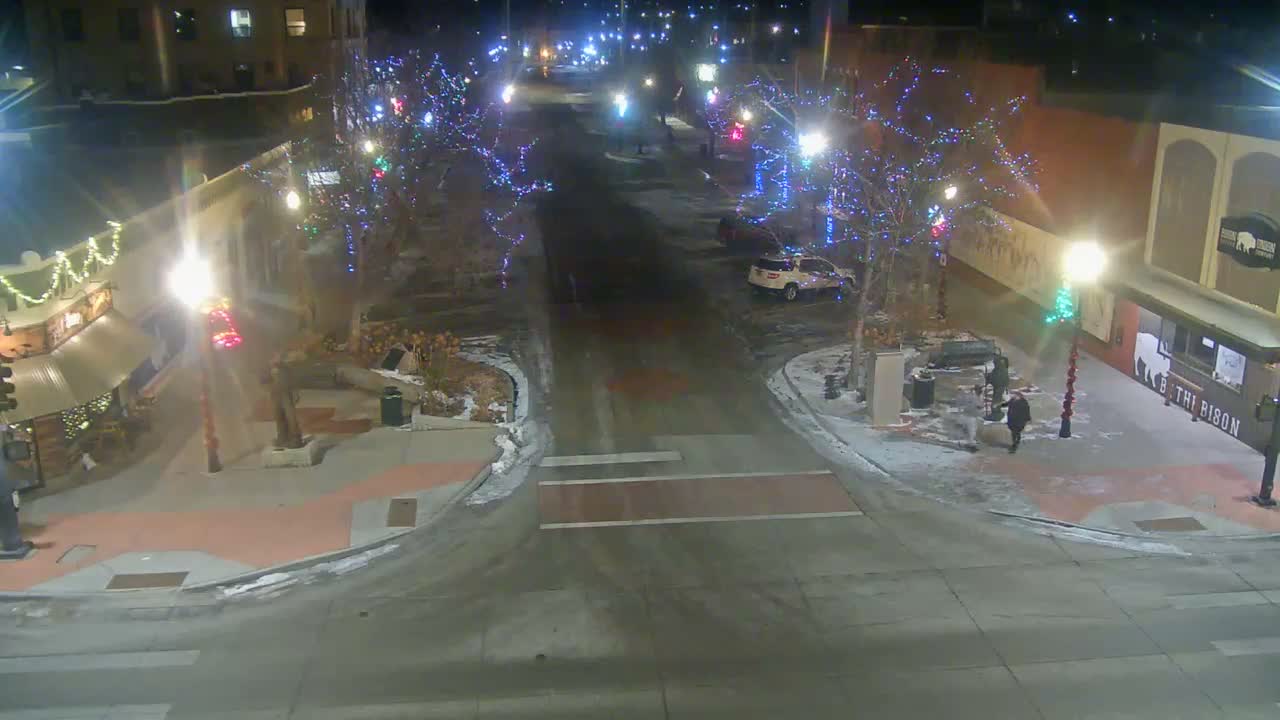 A brightly lit city street at night is adorned with colorful string lights on trees and buildings, with patches of snow covering the ground, suggesting a cold, clear evening, as a white SUV and a lone pedestrian are visible.