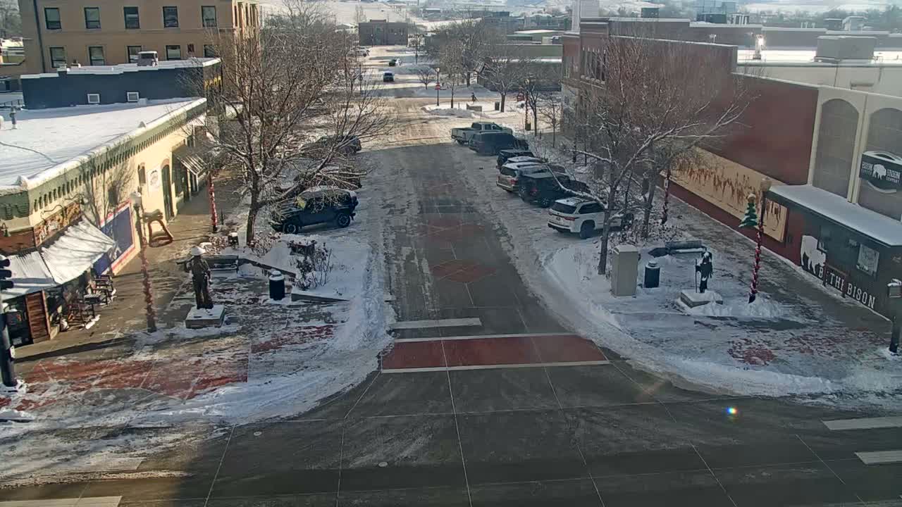 A brightly lit city street at night is adorned with colorful string lights on trees and buildings, with patches of snow covering the ground, suggesting a cold, clear evening, as a white SUV and a lone pedestrian are visible.