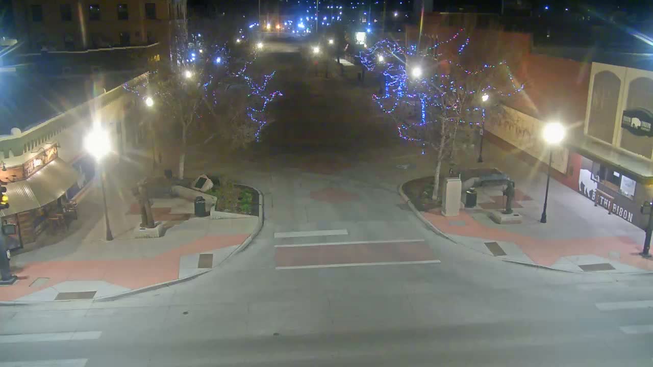 A brightly lit city street at night is adorned with colorful string lights on trees and buildings, with patches of snow covering the ground, suggesting a cold, clear evening, as a white SUV and a lone pedestrian are visible.