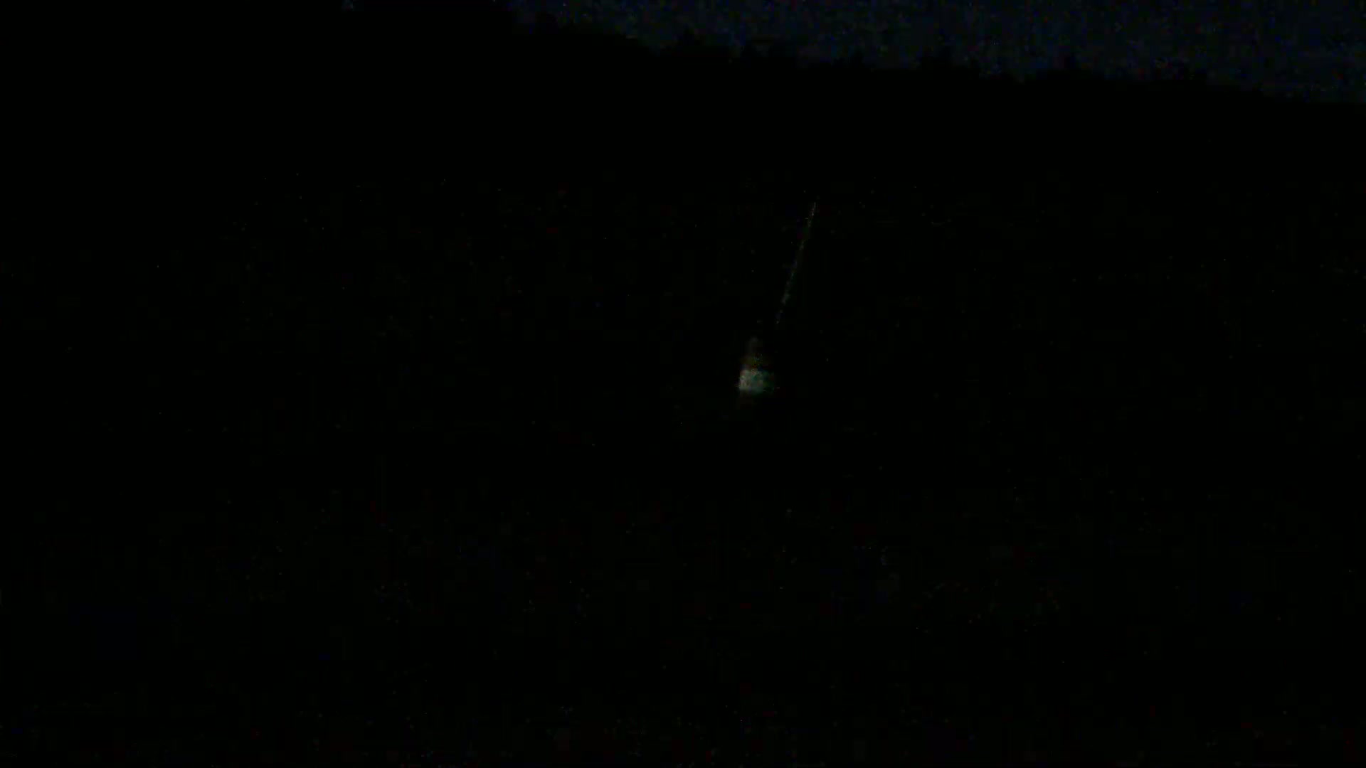 A faintly lit object hangs in the dark night sky above a silhouetted landscape.