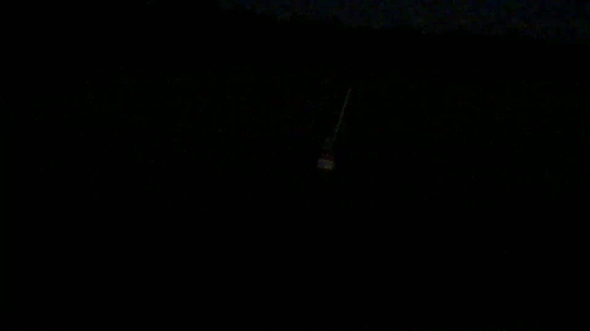 A dimly lit, possibly nighttime scene shows a small, faintly illuminated object hanging from a cable against a dark background.