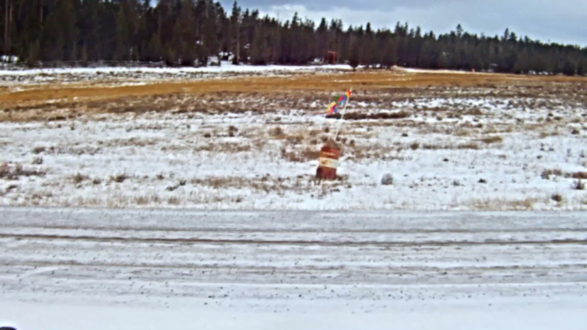 A snowy road borders a field with patches of brown ground, where a striped barrel topped with colorful flags stands before a distant line of evergreen trees, under cloudy and cold weather.
