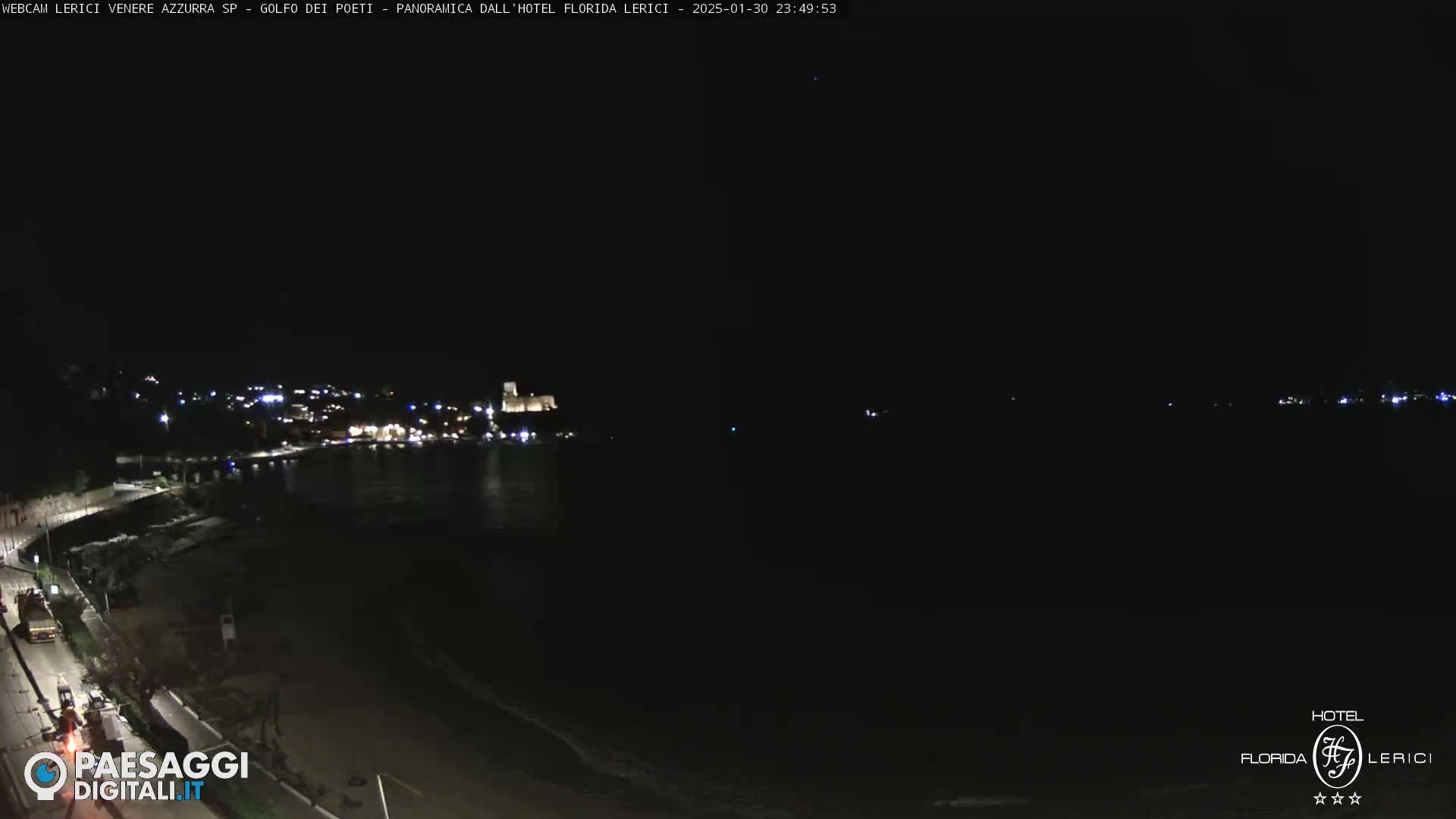 A nighttime coastal scene shows a dark, mostly calm body of water with a dimly lit town and shoreline visible in the distance.