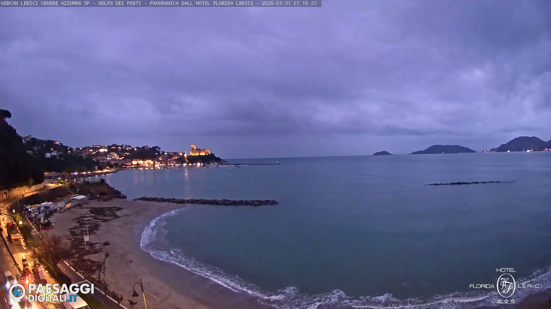 A twilight coastal scene shows a sandy beach, calm sea, and a town with lights nestled against hills under a cloudy sky.