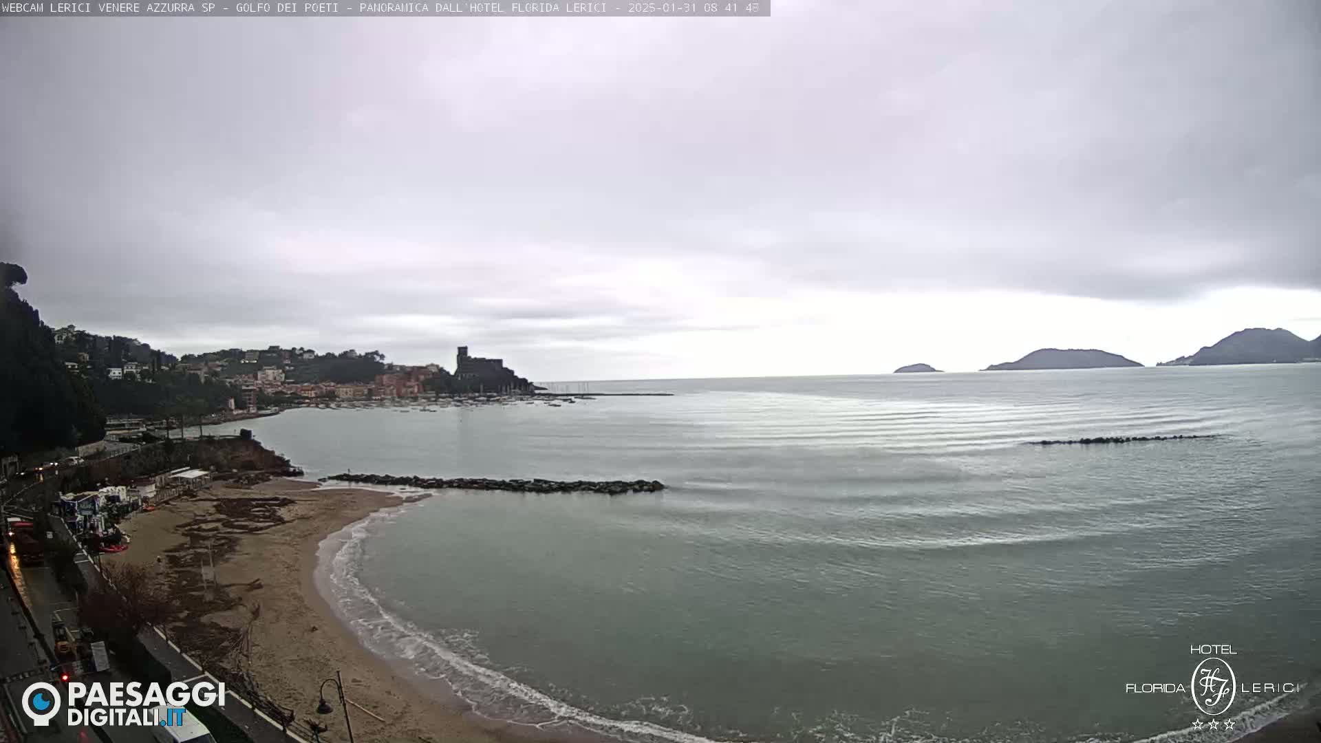 A beach with calm, gray water under an overcast sky, overlooking a town nestled against hills and islands in the distance.
