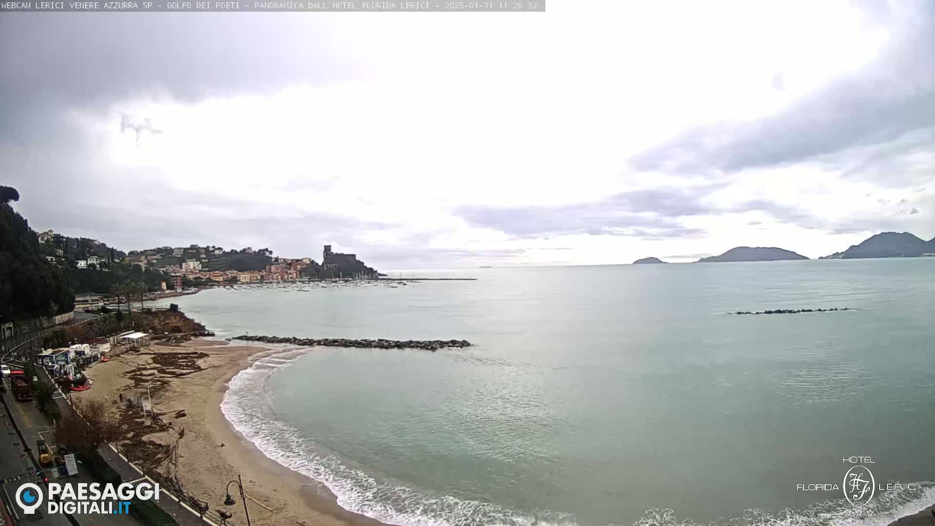 A sandy beach with calm waves and a town nestled beside a calm sea under an overcast sky.