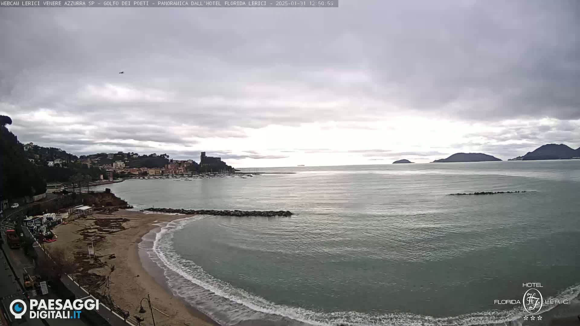 A mostly cloudy day shows a beach with calm waves, a town nestled along a coastline, and islands in the distance.