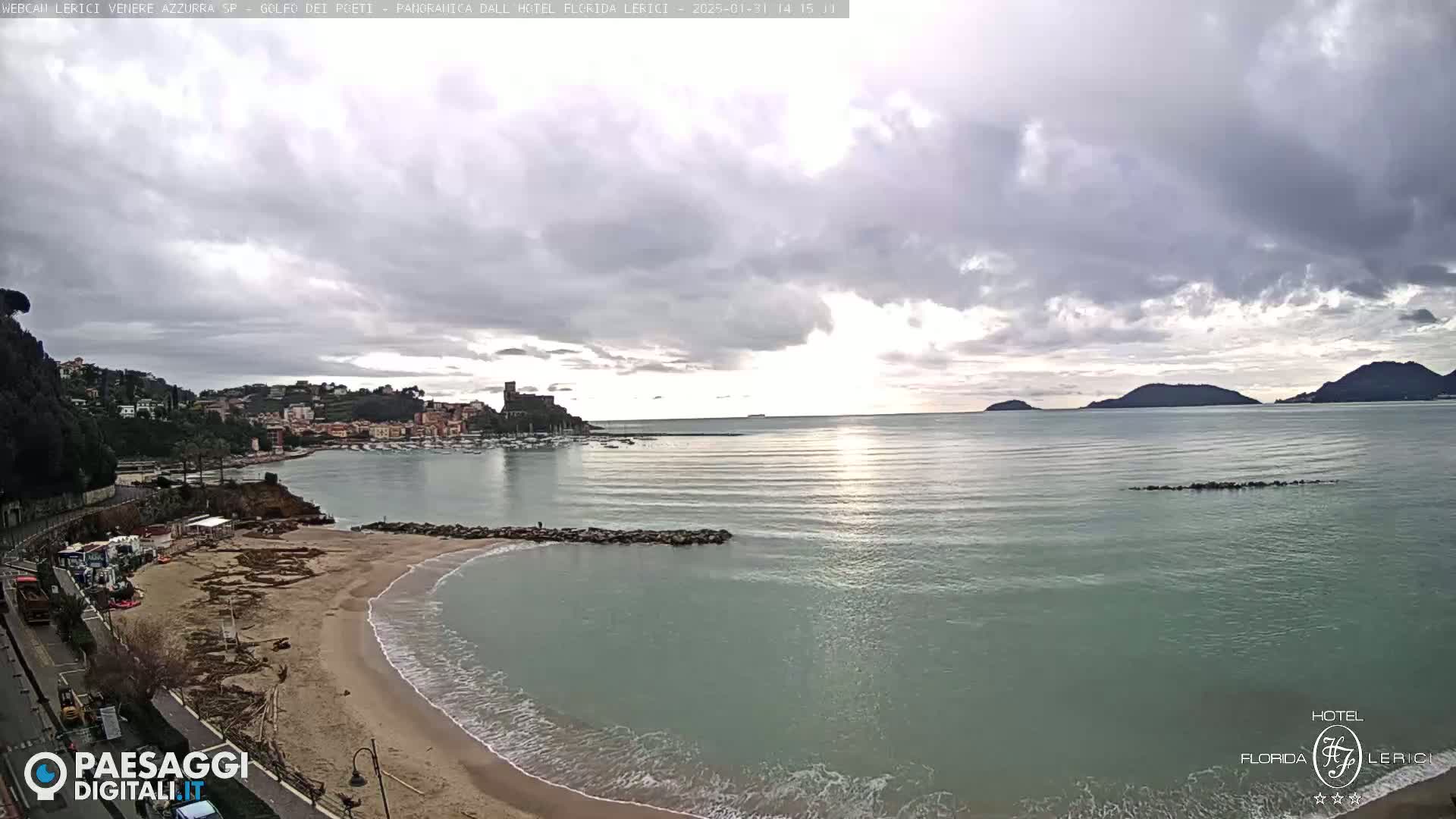 A mostly cloudy day shows a view of a sandy beach and calm sea, with a town and distant hills visible in the background.