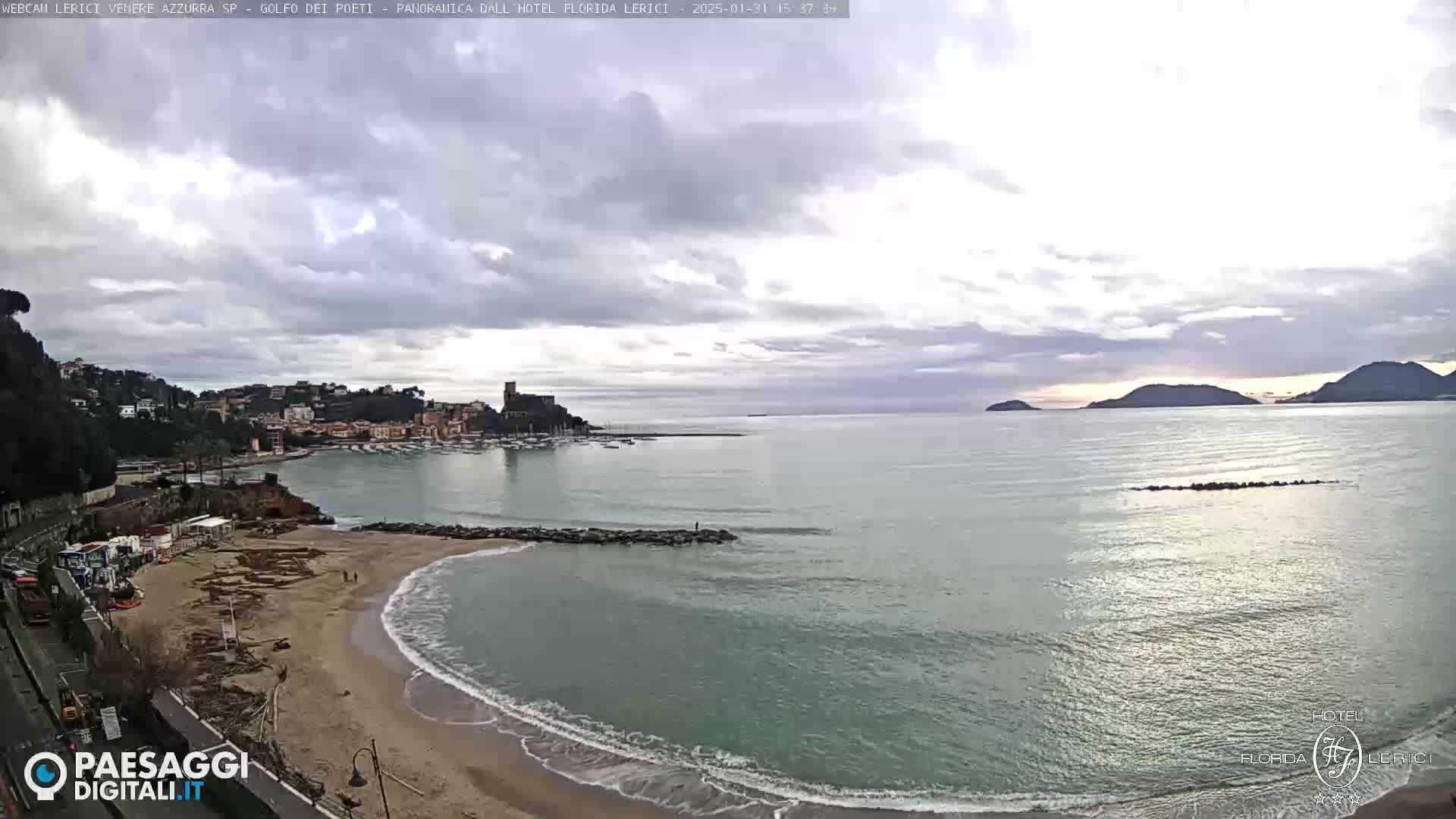 A coastal town with a sandy beach and calm sea is seen under a mostly cloudy sky.