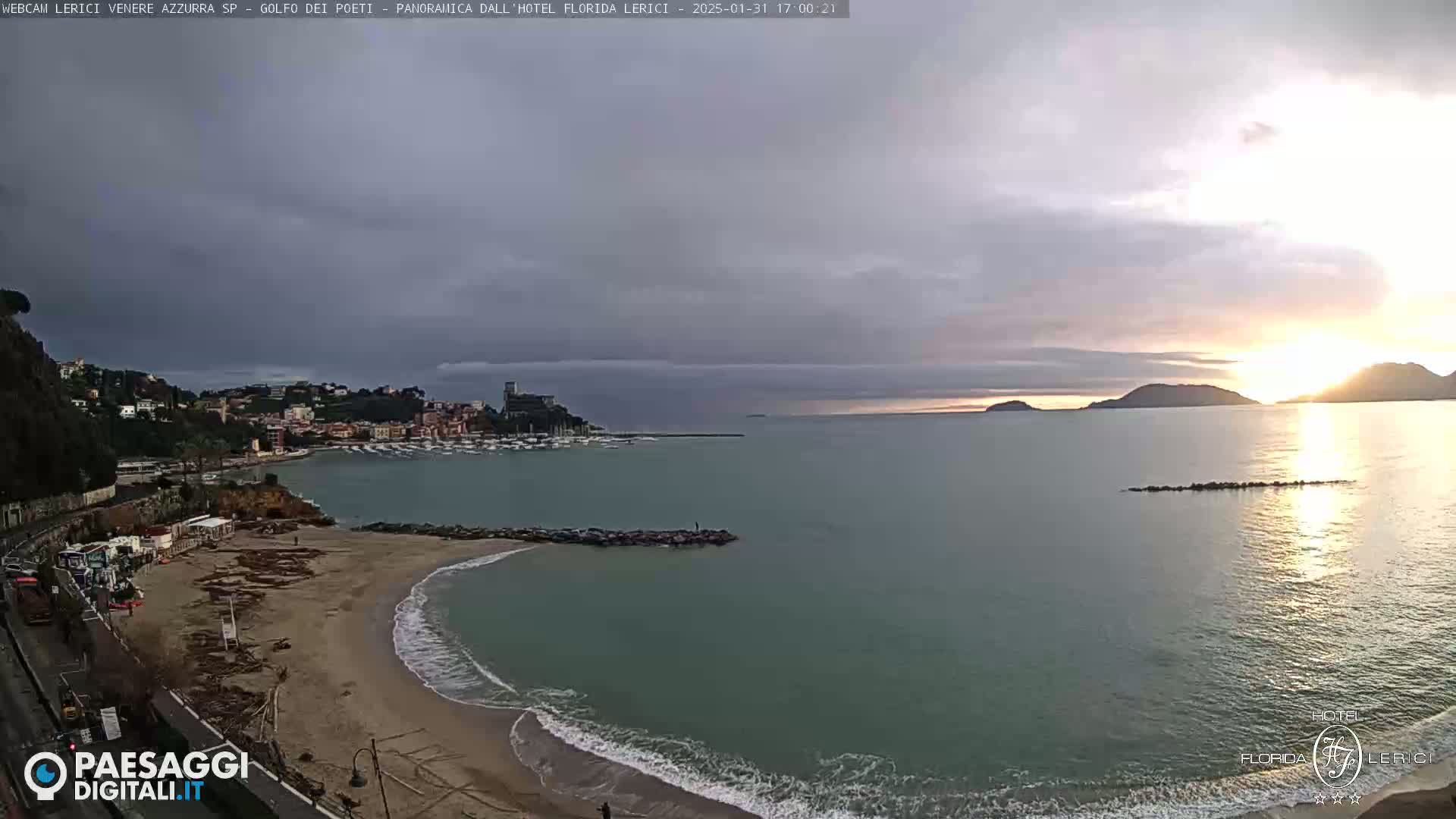 A coastal town with a sandy beach and calm sea is seen at sunset under a mostly cloudy sky.