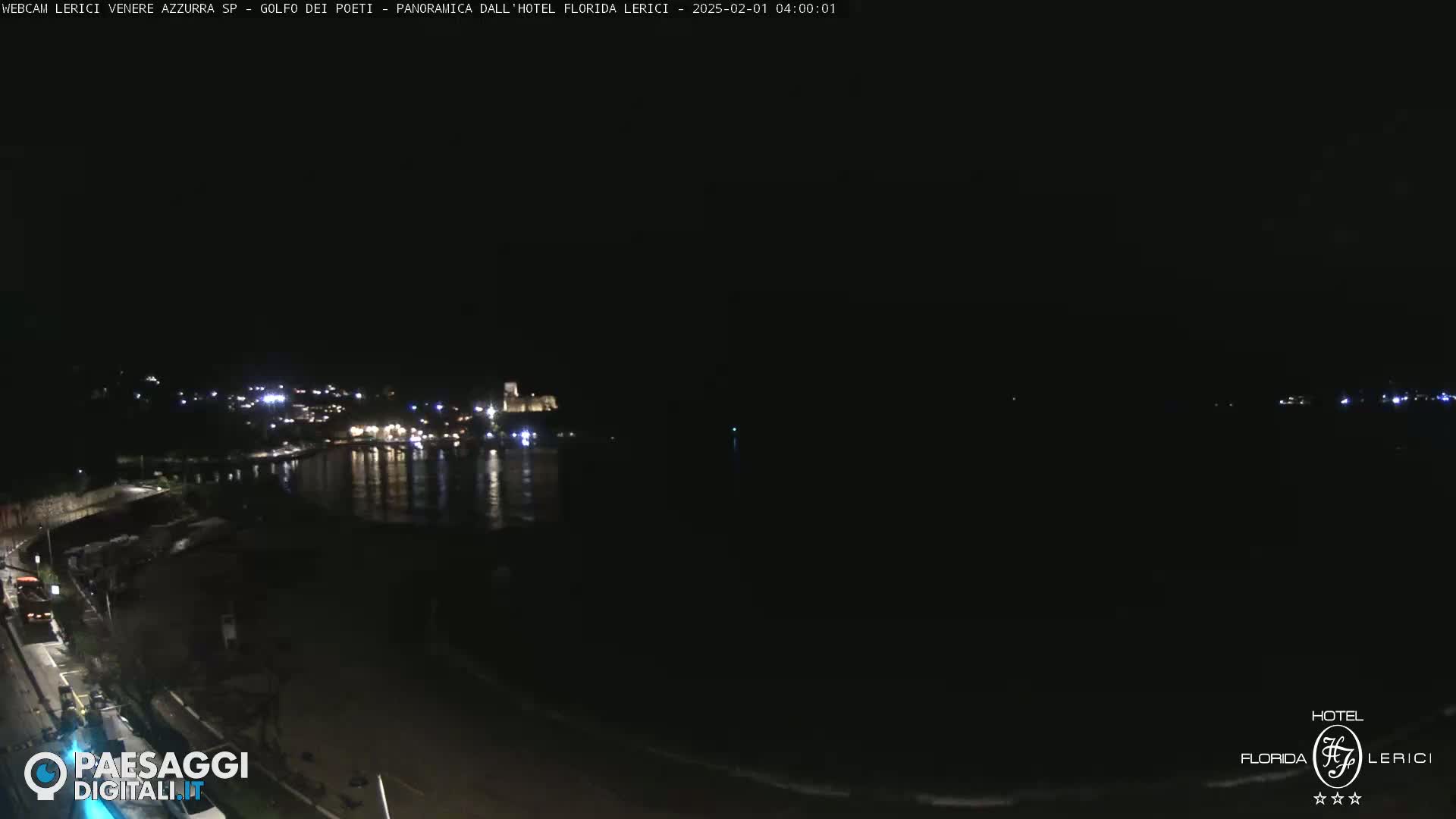 A nighttime, seaside view shows a dark beach, calm water reflecting dim lights from a town across the bay, under a clear, dark sky.