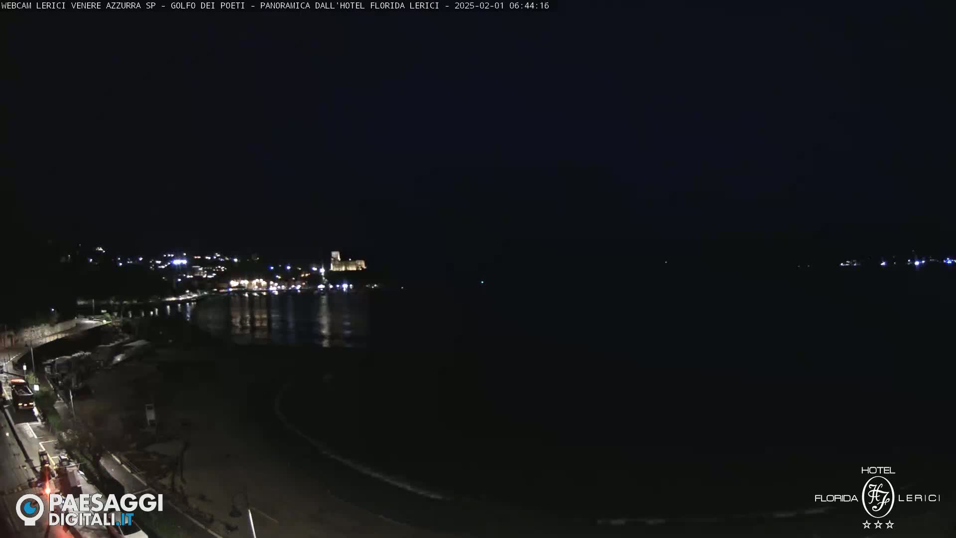 A nighttime view of a dark, calm body of water next to a dimly lit shoreline with buildings visible in the distance.