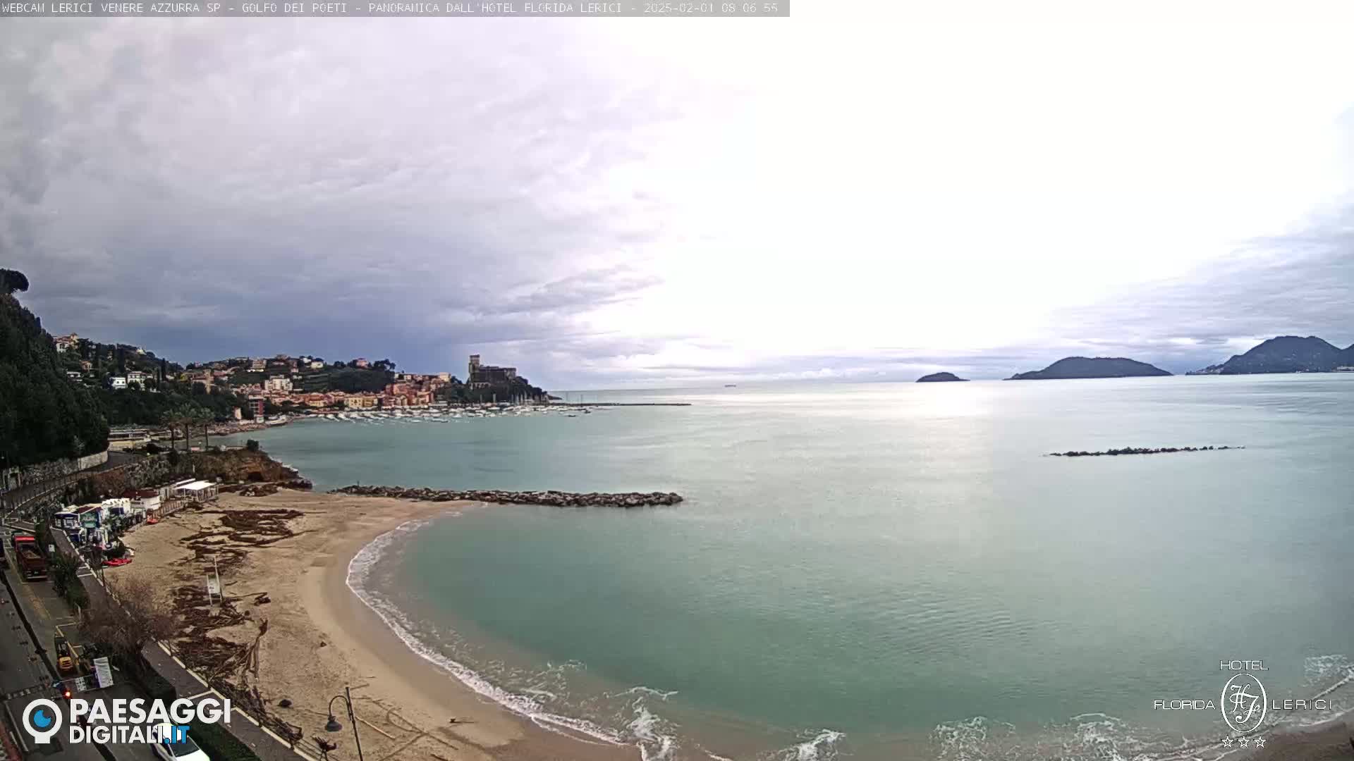 A sandy beach curves into a calm, teal sea under a cloudy sky, with a town and distant mountains visible in the background.