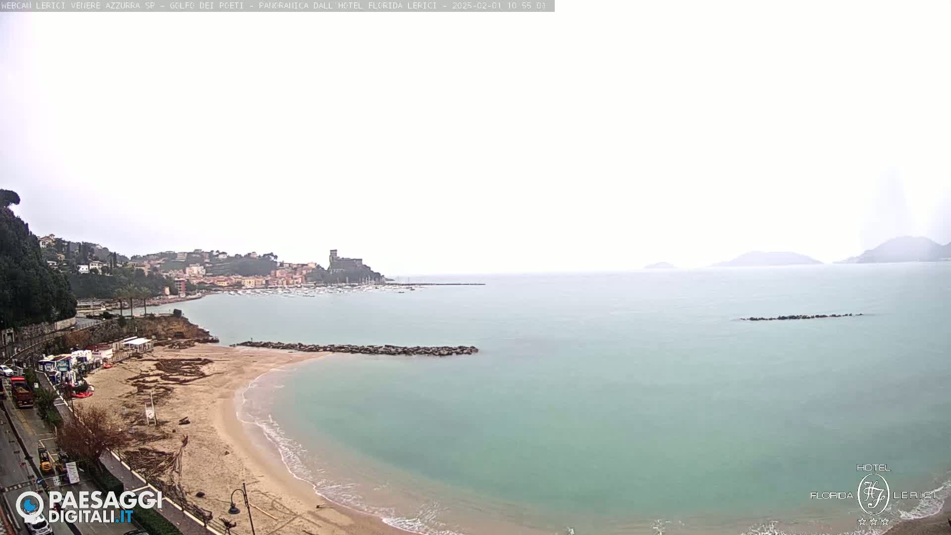 A sandy beach curves into a calm, light-green sea under an overcast sky, with a town and hills visible in the background.