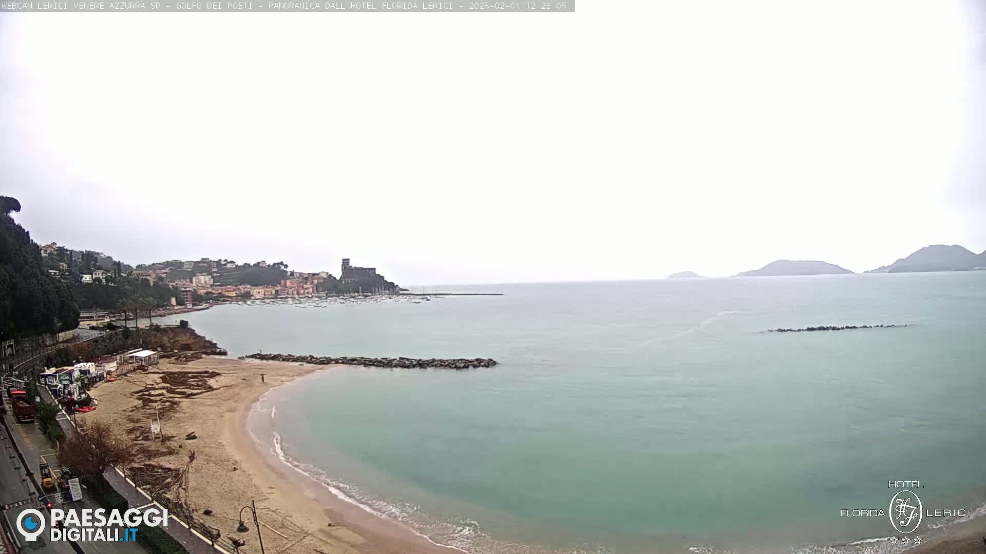 A beach with calm, light-green water and a town in the background under an overcast sky.