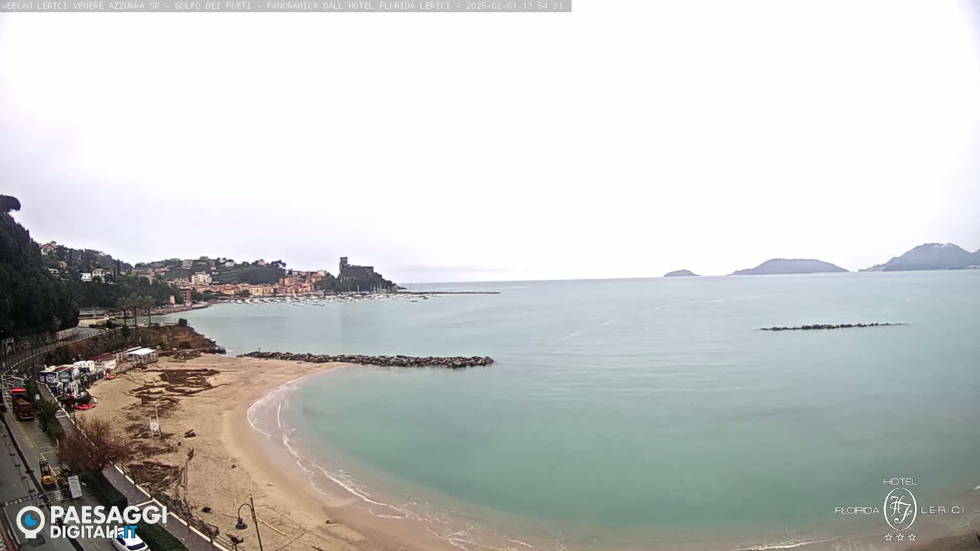 A sandy beach curves into a calm, light-green sea under an overcast sky, with a town and distant hills visible across the water.