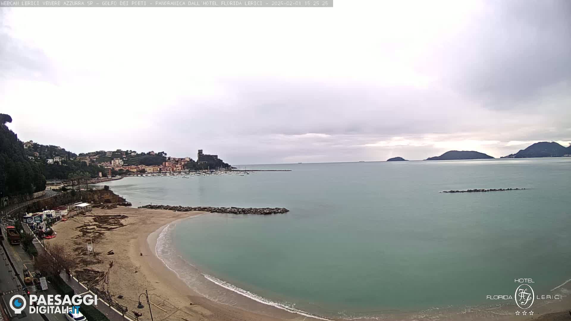A sandy beach curves into a calm, teal-colored sea under an overcast sky, with a town and distant hills visible across the bay.