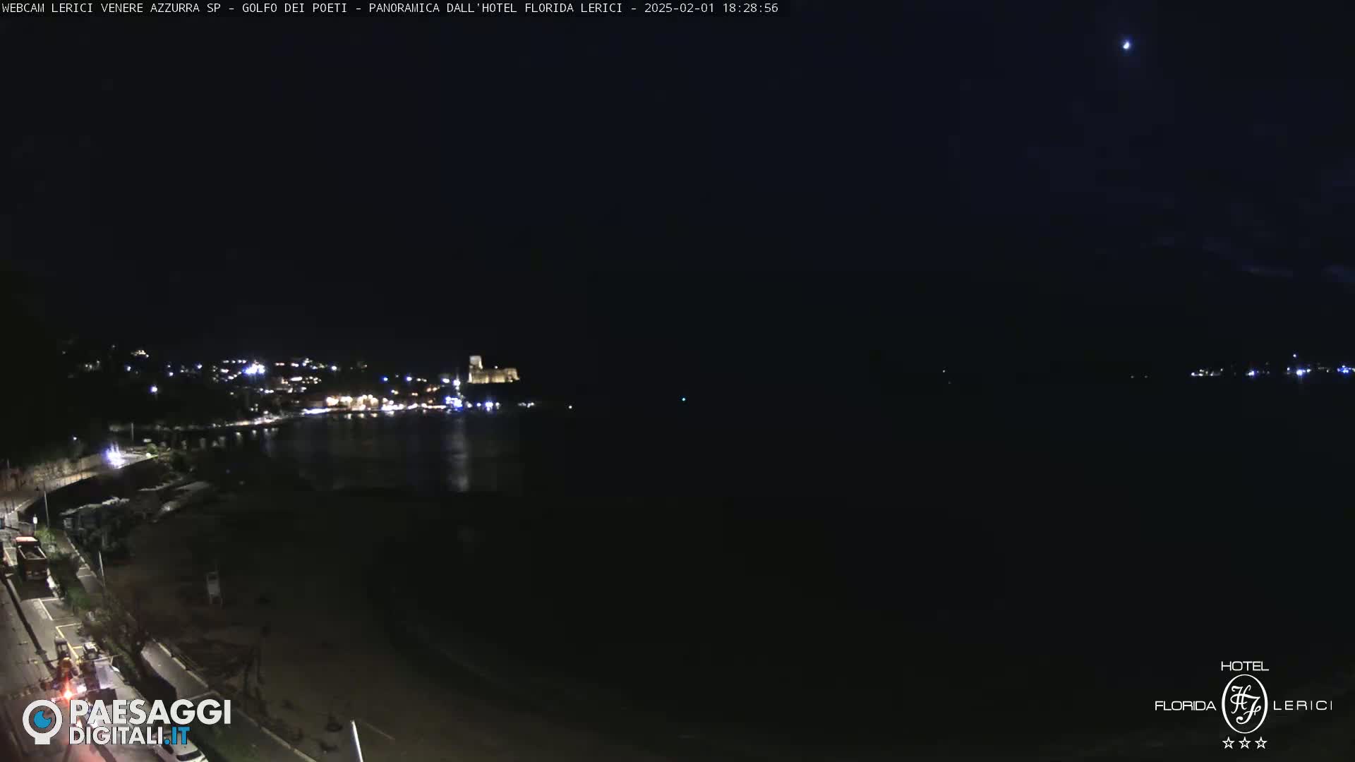 A nighttime coastal scene shows a dark body of water reflecting the lights of a town across the shore, under a clear, dark sky.
