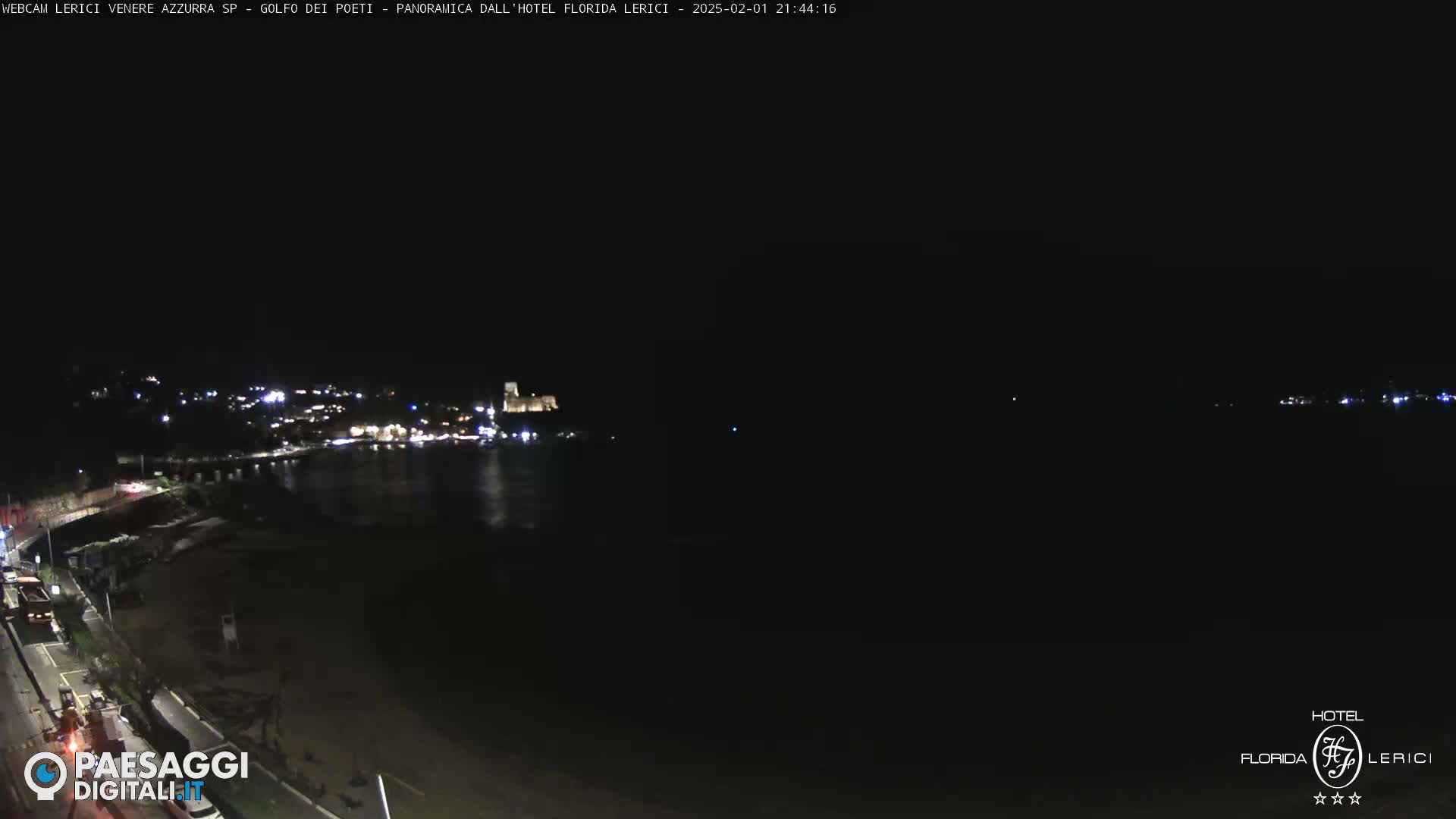 A nighttime coastal scene shows a dark beach and calm water, with a town's lights visible in the distance across the water.