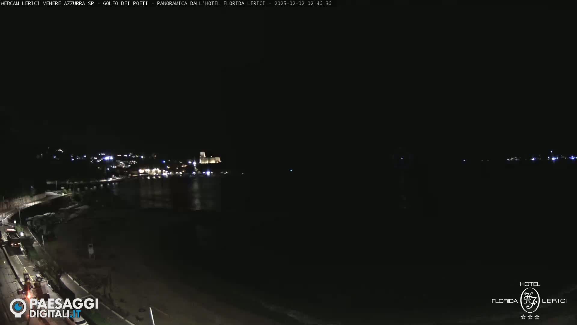 A nighttime coastal scene shows a dark beach and calm water with a town's lights visible in the distance.
