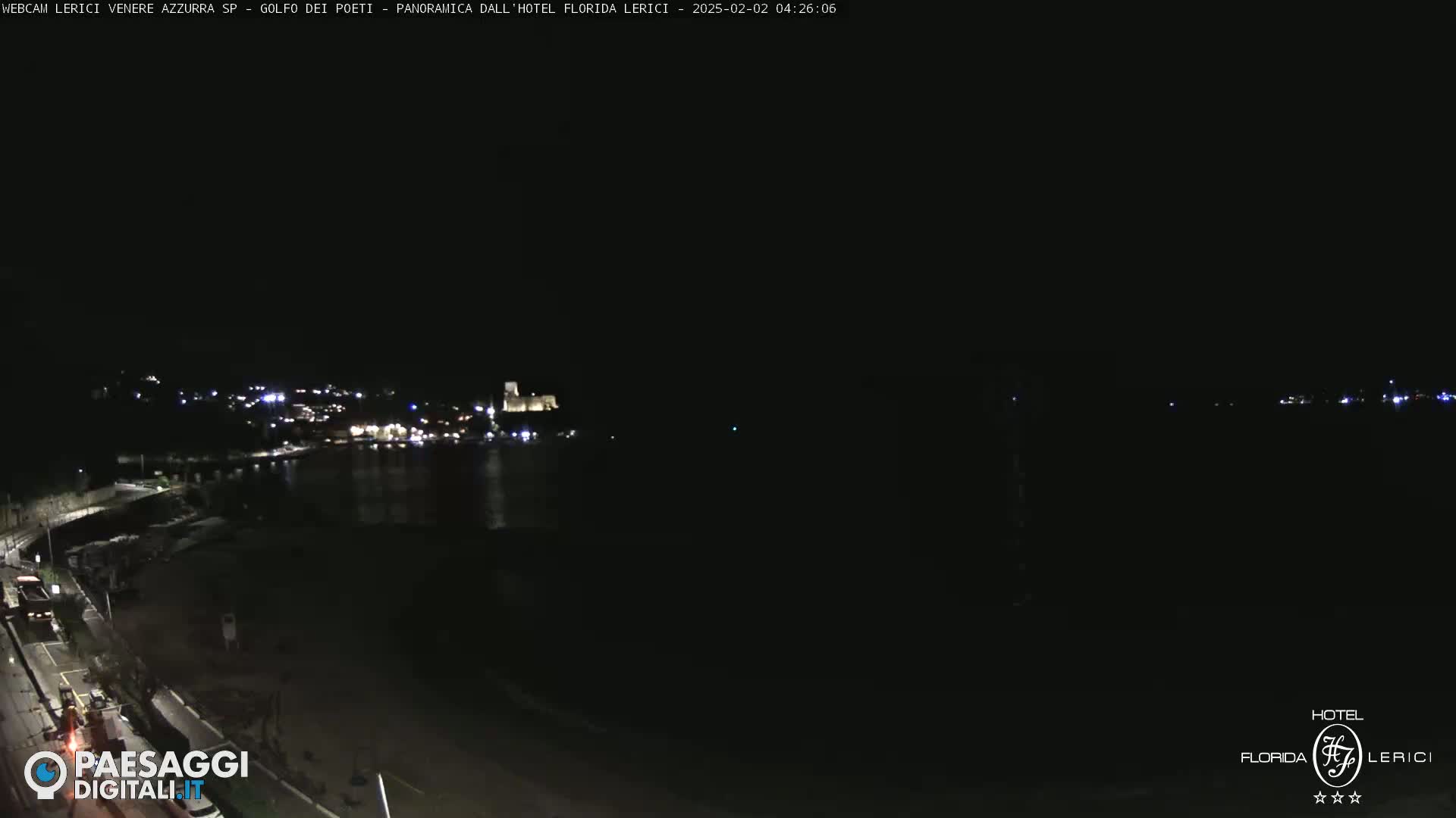 A nighttime view of a dark coastline with a town's lights faintly visible across a body of water.