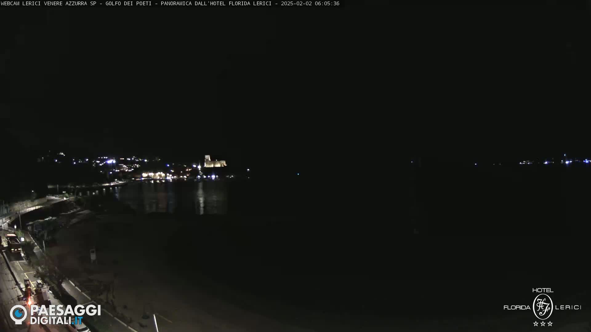 A nighttime coastal scene shows a dark body of water reflecting faint lights from a town nestled on a hillside.