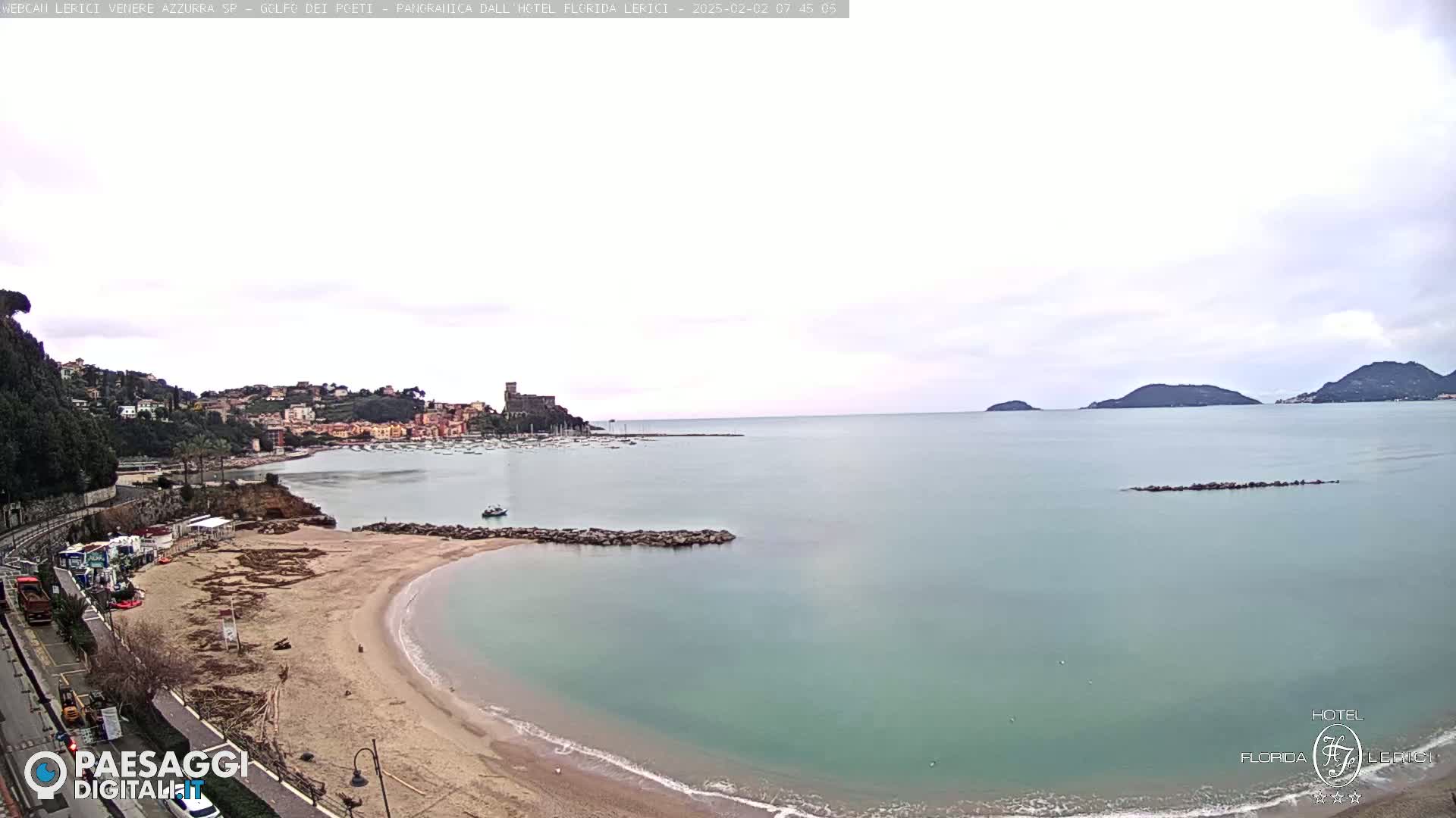 A mostly cloudy day reveals a sandy beach curving into a calm, teal-green sea, with a town and distant islands visible in the background.