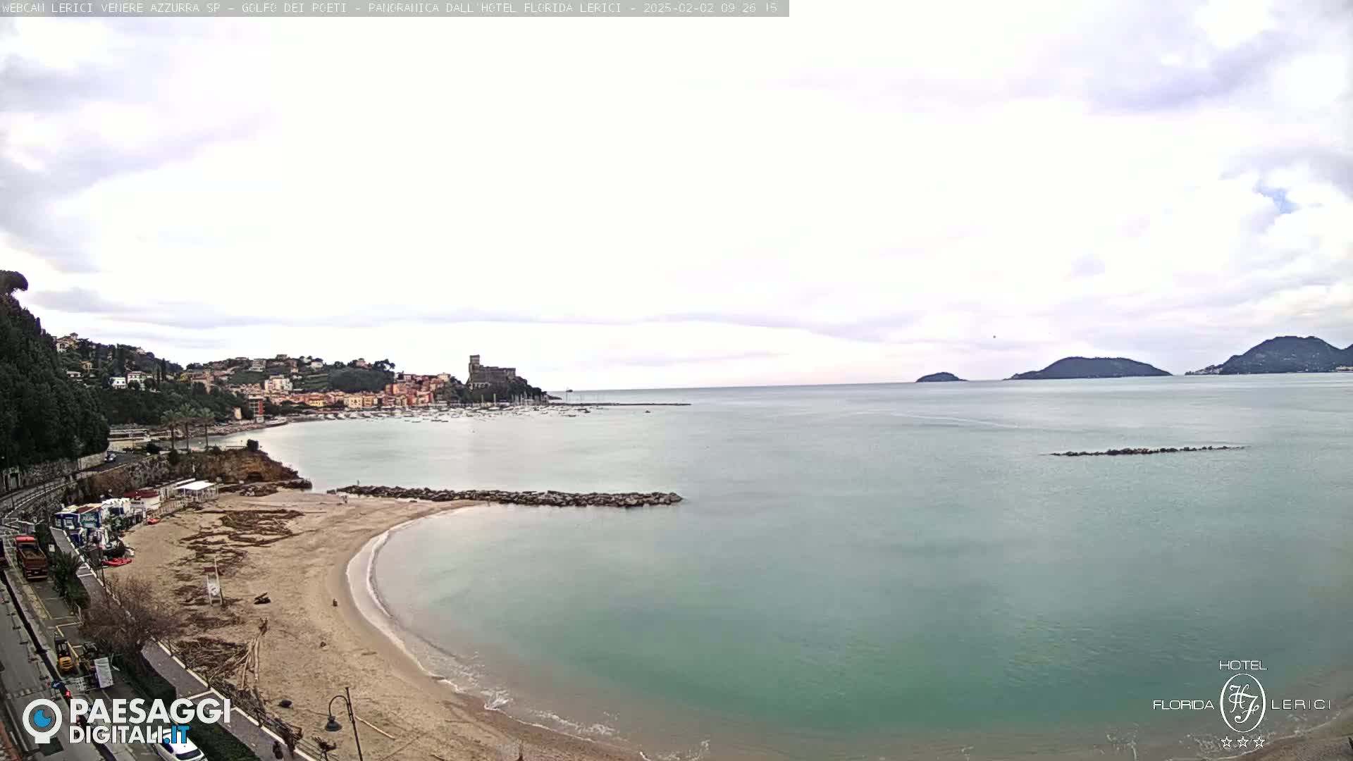 A mostly cloudy day reveals a sandy beach curving into a calm teal sea, with a town and distant hills visible across the bay.