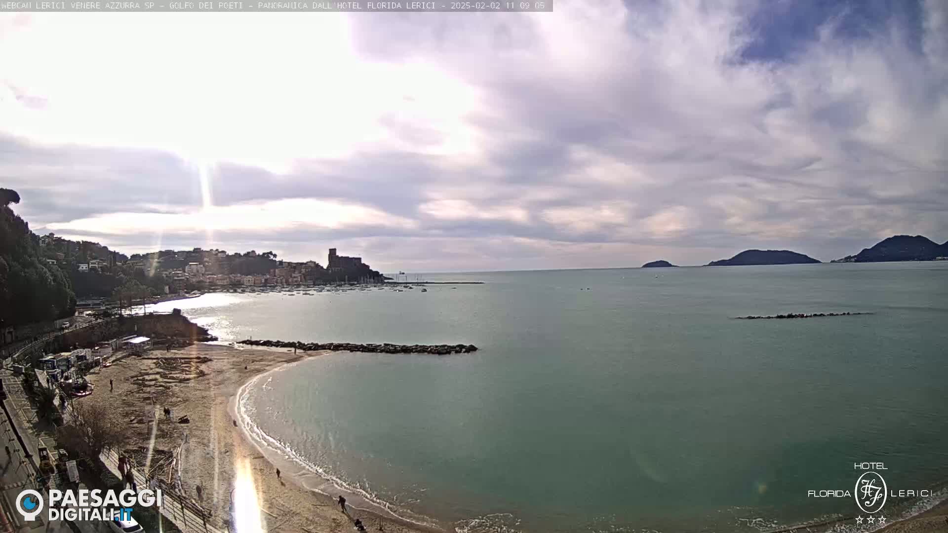 A partly cloudy day reveals a sandy beach curving into a calm bay, with a town and a castle visible in the distance across the water.