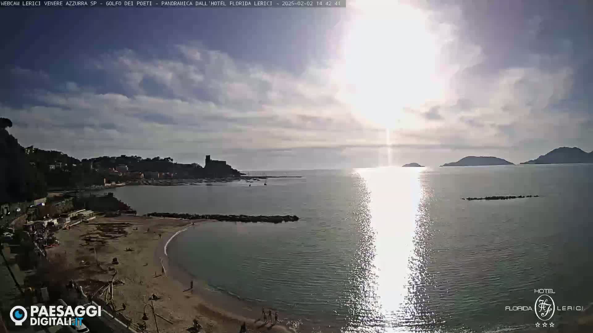 A sunny day reveals a sandy beach curving into a calm bay, with a town and distant hills visible across the water.