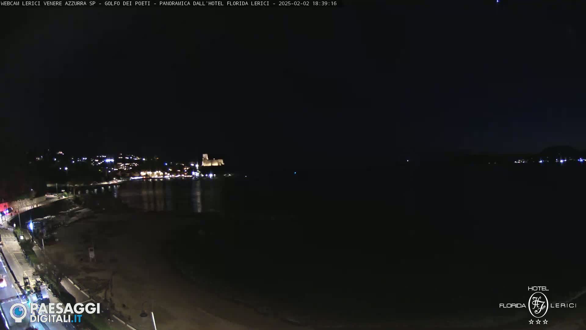 A nighttime, coastal scene shows a dark sandy beach, a calm body of water, and a town with lights across the water under a clear, dark sky.